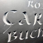 Closeup detail of letter carving. The image shows a fishing hook hanging from the capital letter C. Relief in stone.