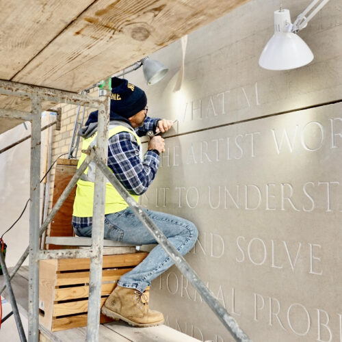 Large limestone wall at Raclin-Murphy Museum of Art | University of Notre Dame. Javier R.Alfonzo is sitting on a scaffolding while carving large letters on the wall.