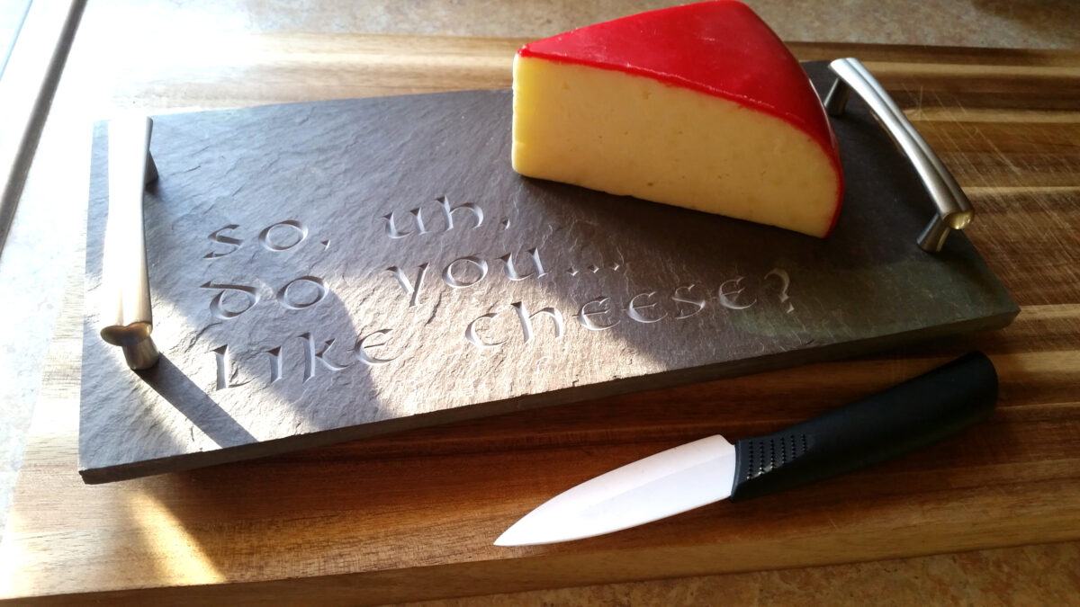 An image of a purple slate cheese tray with a waxed covered chunk of gouda cheese and the inscription "So.. do you like cheese?, carved. 
